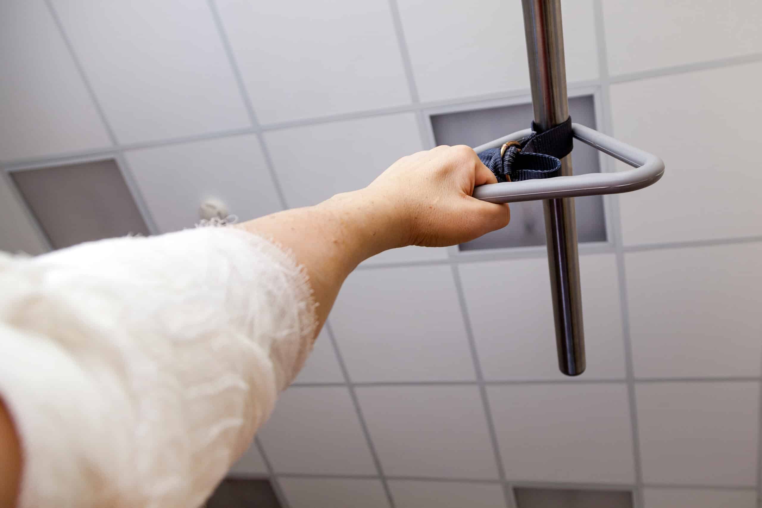 Main tenant une poignée de barre de traction triangulaire fixée à une barre métallique, bras bandé sous une couverture blanche, plafond de dalles.