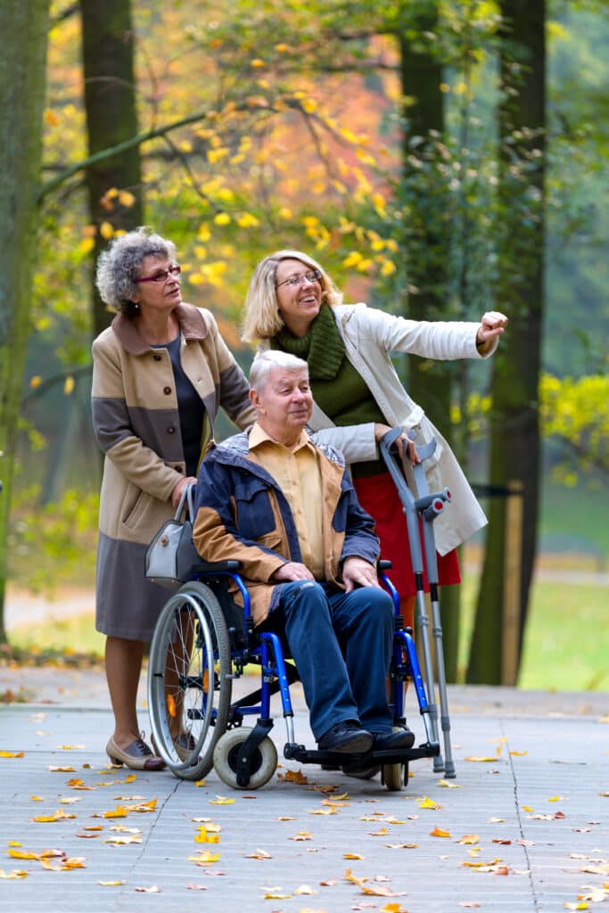 Personne âgée en fauteuil roulant avec deux femmes, une désignant quelque chose au loin, dans un parc d'automne.